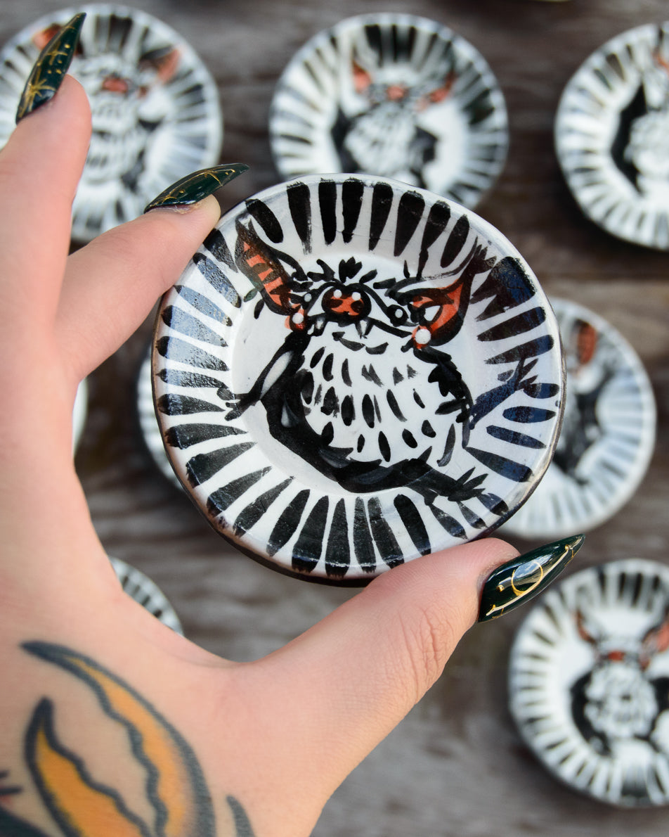 Hand holding a small ceramic dish with a painted design of a bat surrounded by similar dishes on a wooden surface.