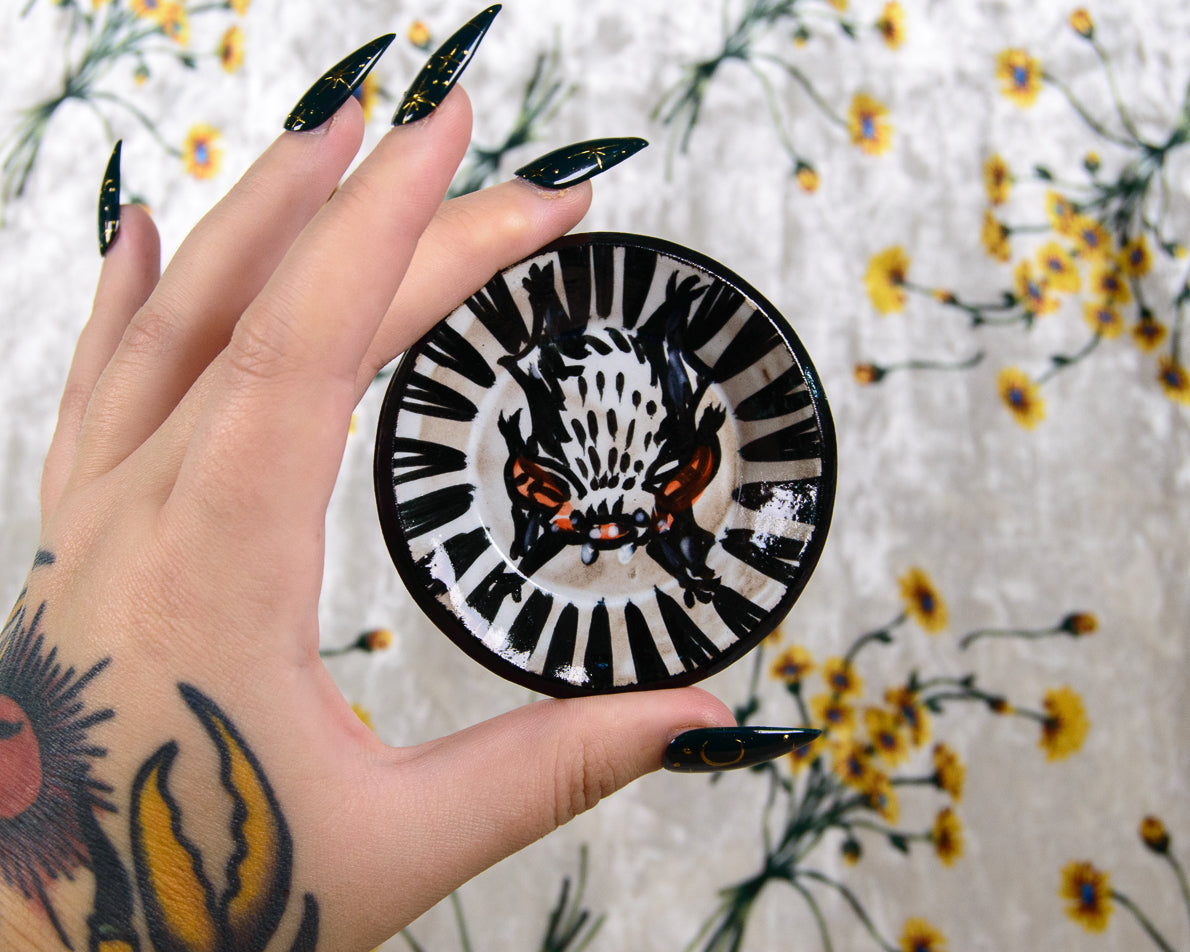 Hand holding a round black and white ceramic dish with a bat design against a floral background