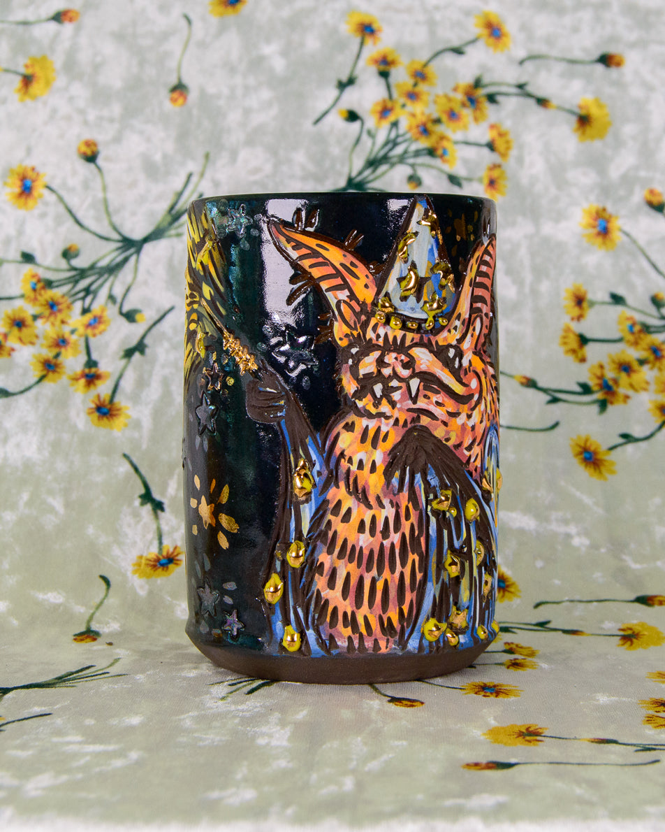 Black stoneware cup with an orange and blue cartoon wizard bat design and gold luster stars