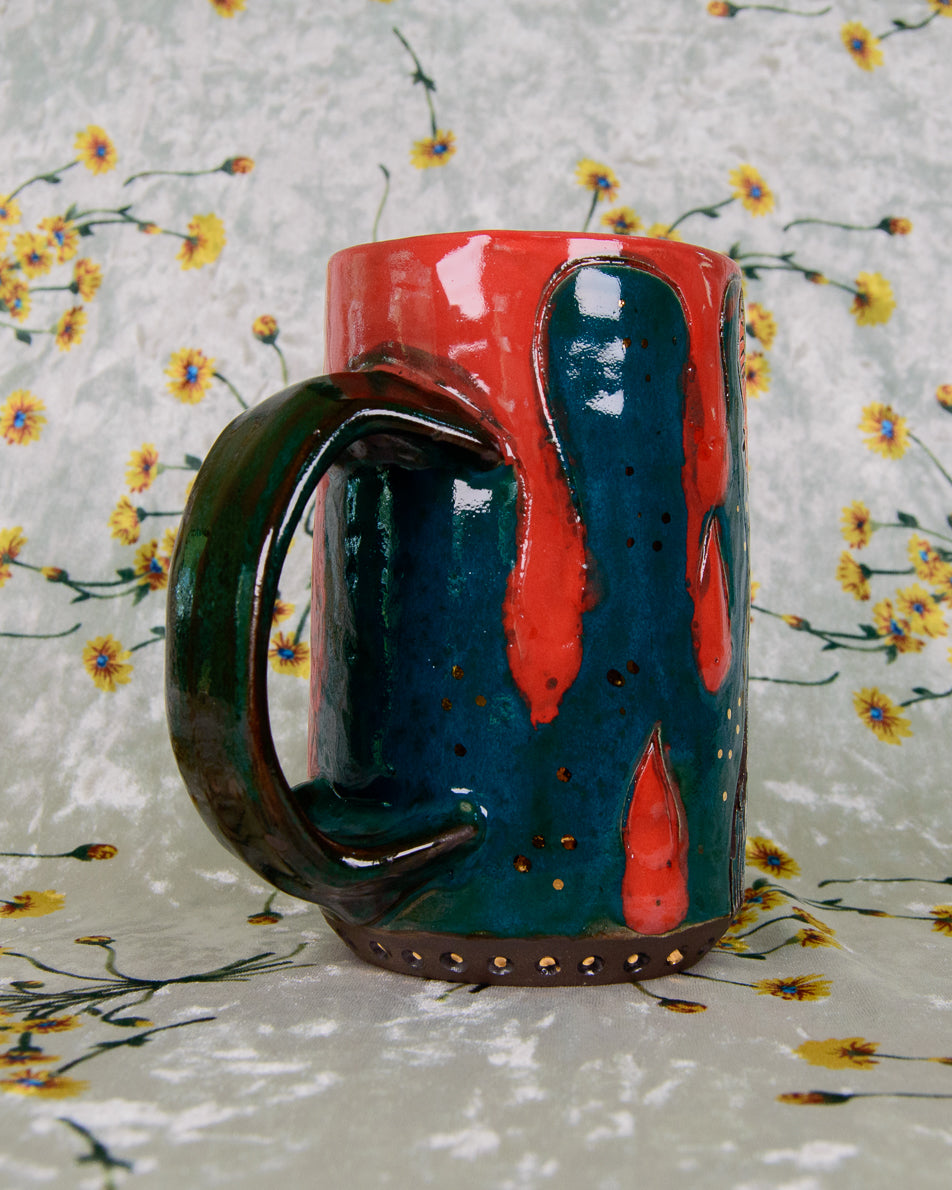 Blue Ceramic mug with bright red blood drip designs and gold accents on a floral-patterned fabric background