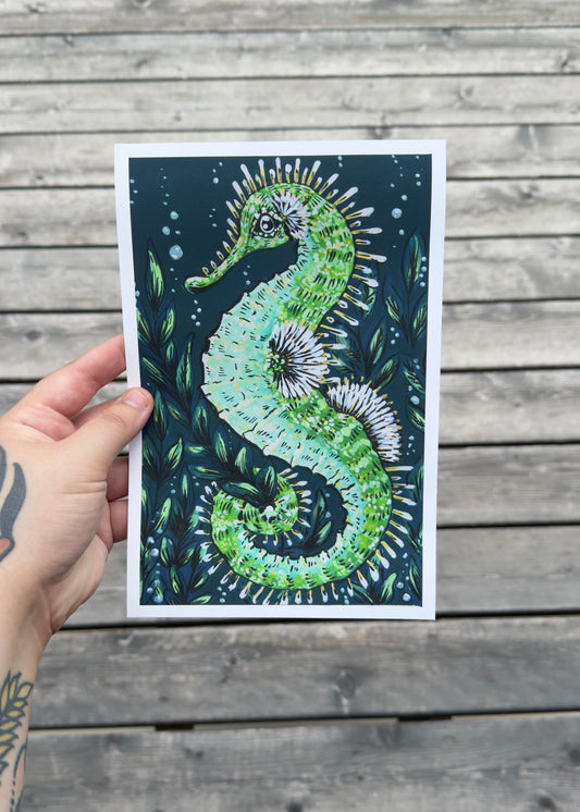 Hand holding a card with a green seahorse design on a wooden surface