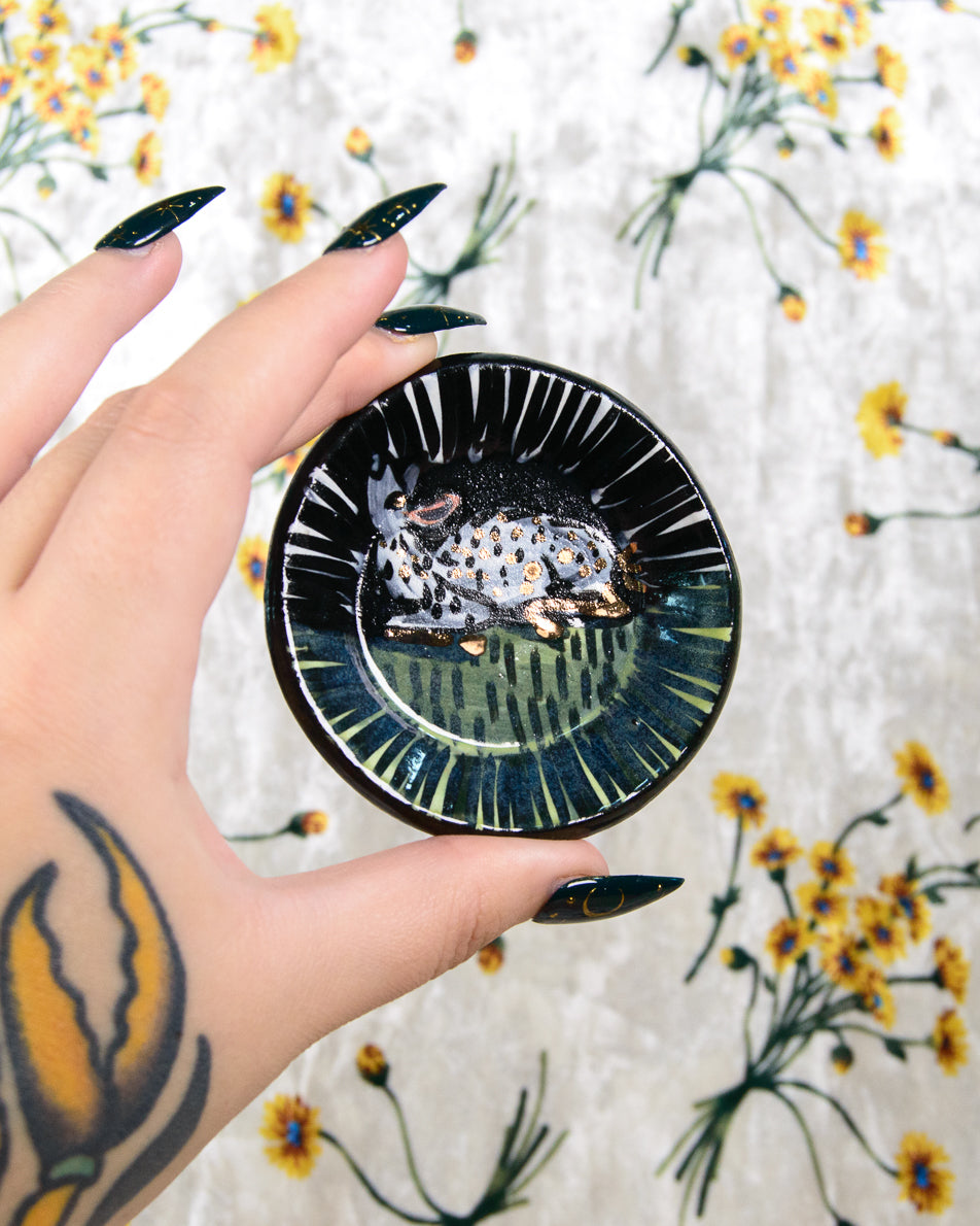 Hand holding a black ceramic dish with a white fawn design on a floral-patterned fabric background