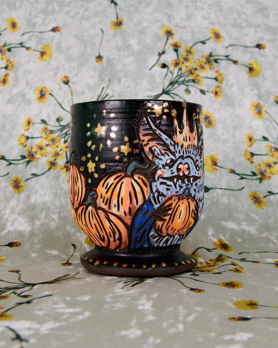 Black stoneware cup with a blue bat design and pumpkins on a matte black background with gold luster stars