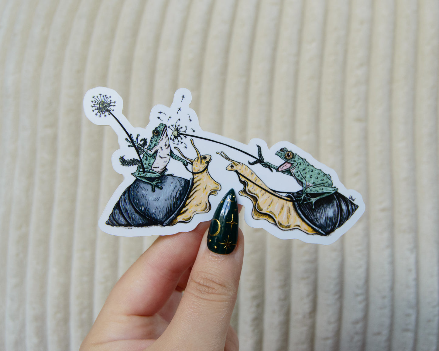 Hand holding a sticker with two frogs riding snails and jousting with dandelions against a beige background