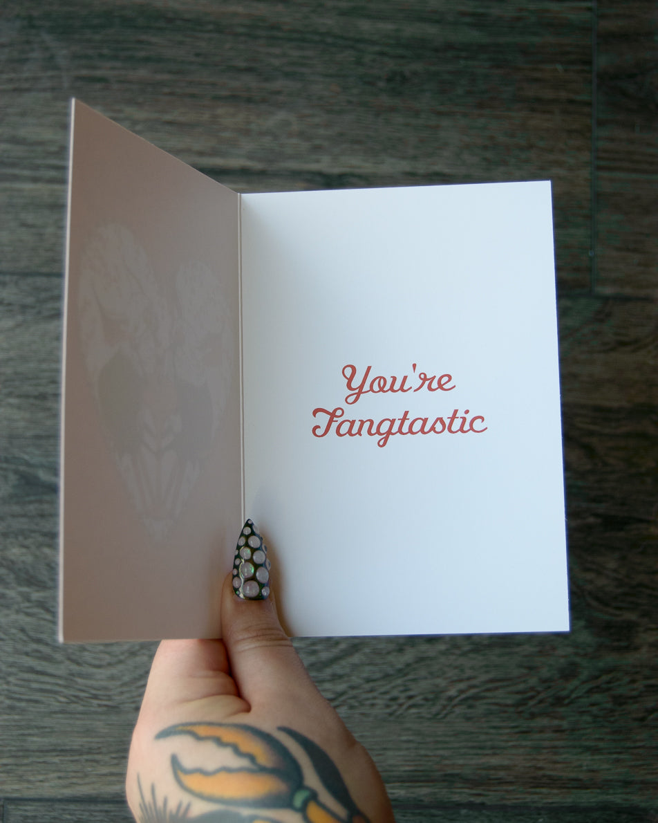 Hand holding a card with 'You're Fangtastic' text on a wooden surface