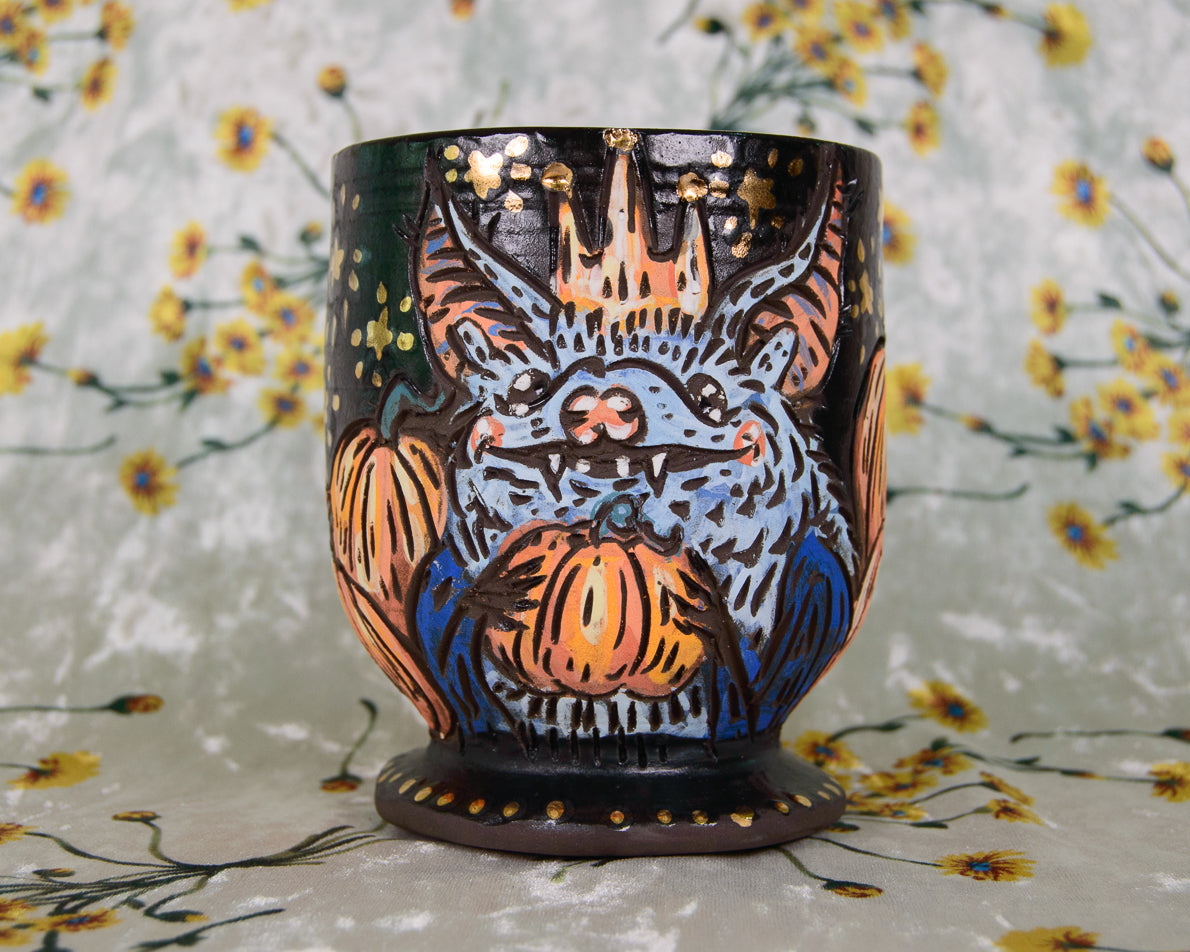 Ceramic goblet cup with a blue bat design holding pumpkins on a matte black background. Decorated with shiny gold stars.