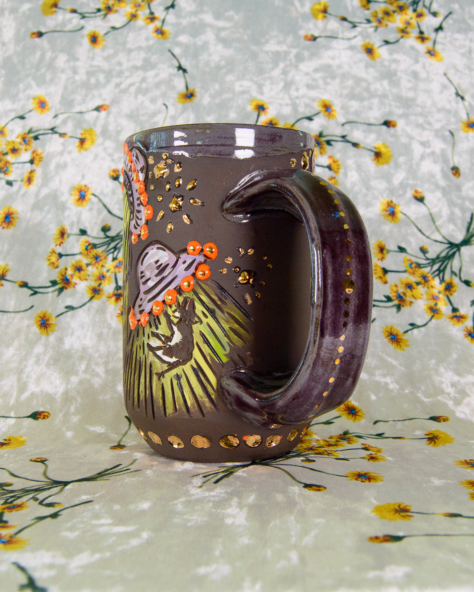 black ceramic mug with UFO and cow designs, purple and gold handle