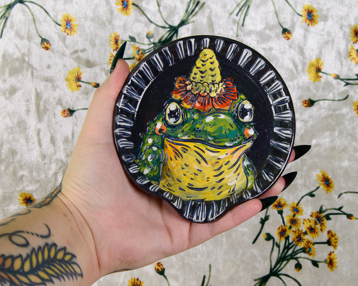 Hand holding a decorative plate with a colorful frog design against a floral background