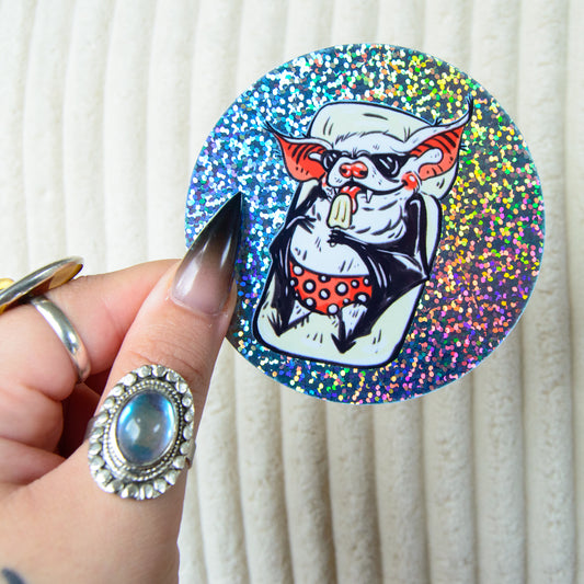 Glittery round sticker of a bat lounging in a pool wearing sunglasses and licking a popsicle