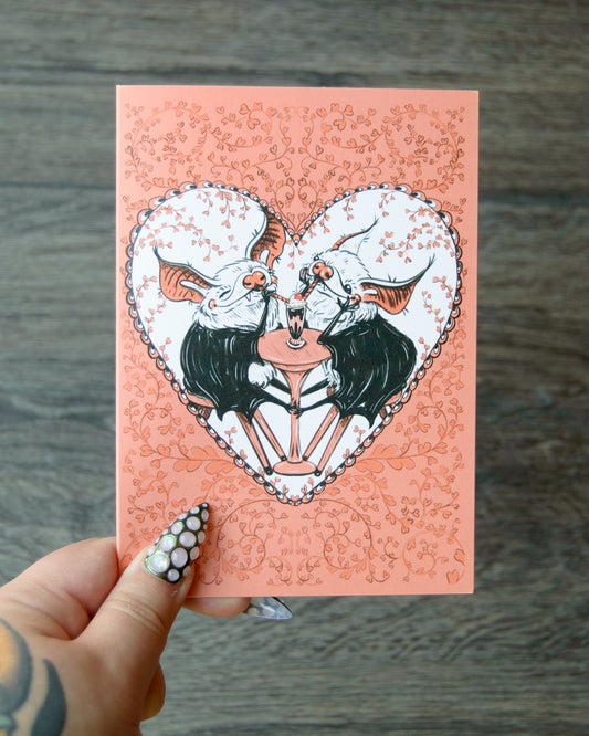 Card with a heart design featuring two bats in love on a pink background, held by a hand.