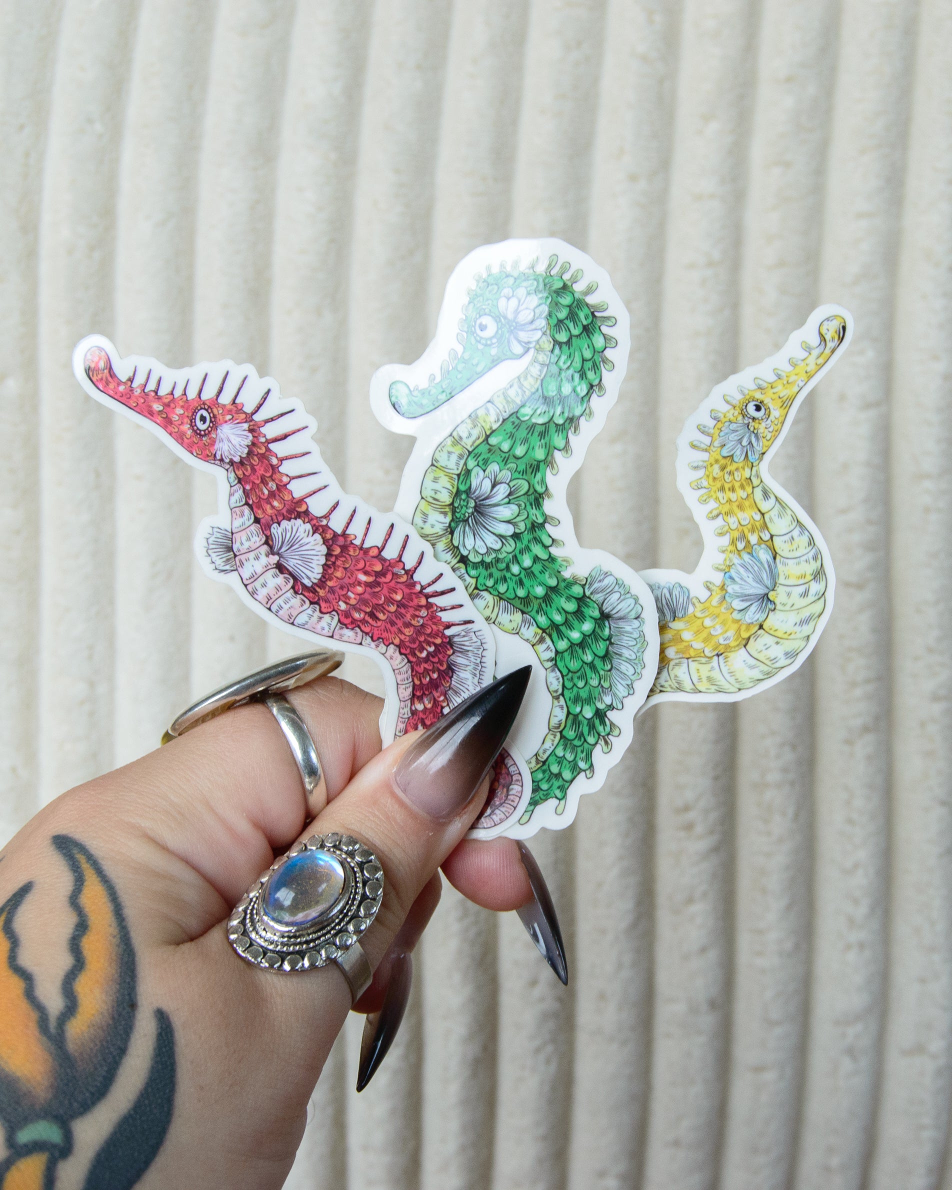 Stickers of Colorful Illustrations of seahorses held by a hand with tattoos and rings against a neutral background.