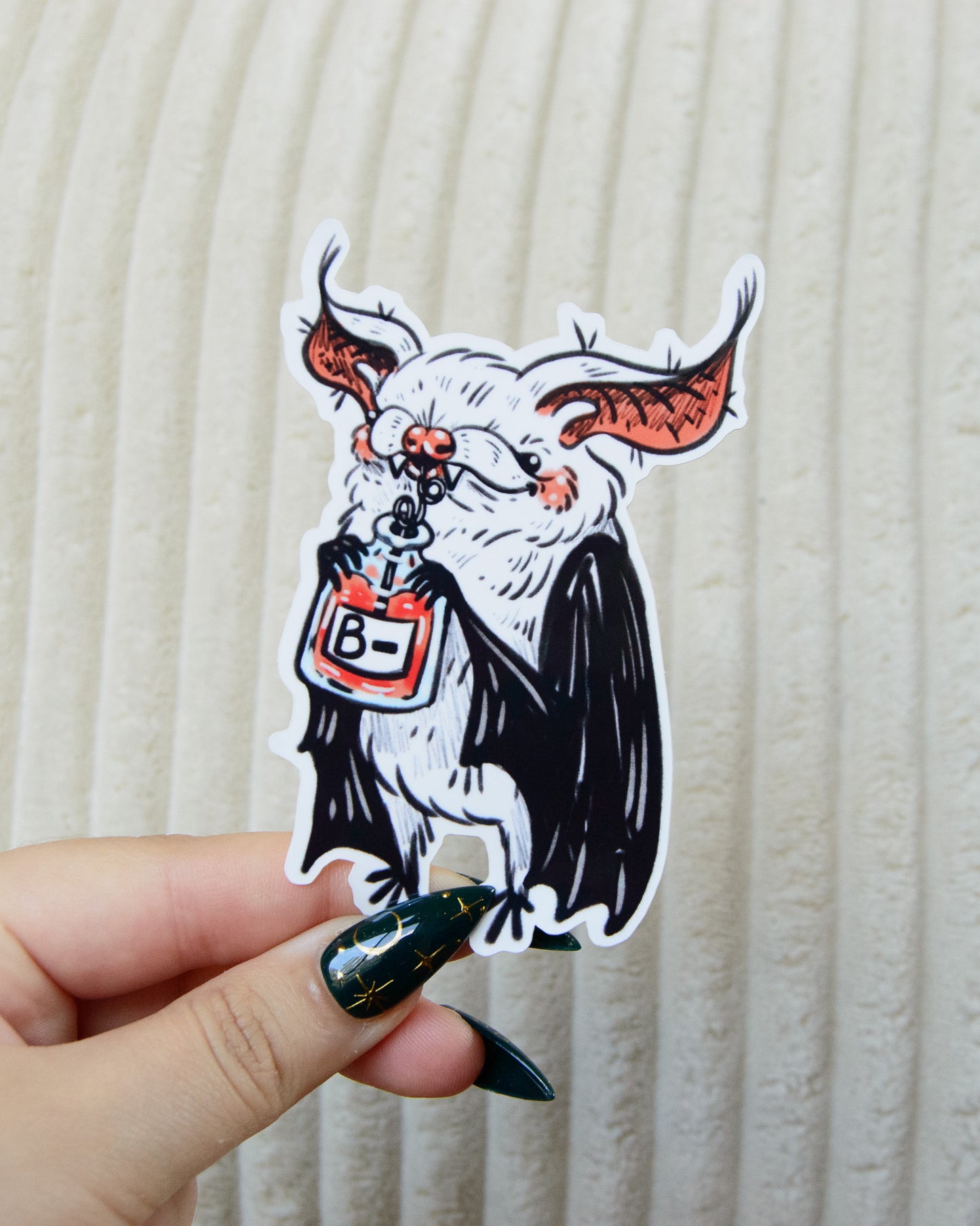 Sticker of a stylized bat drinking a blood bag labeled 'B-'l held by a hand against a beige background