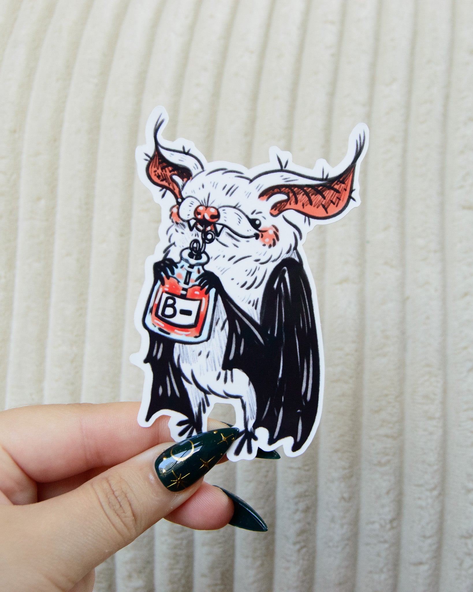 Sticker of a stylized bat drinking a blood bag labeled 'B-'l held by a hand against a beige background