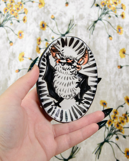 Hand holding a round ring dish with a bat design on a floral-patterned fabric background