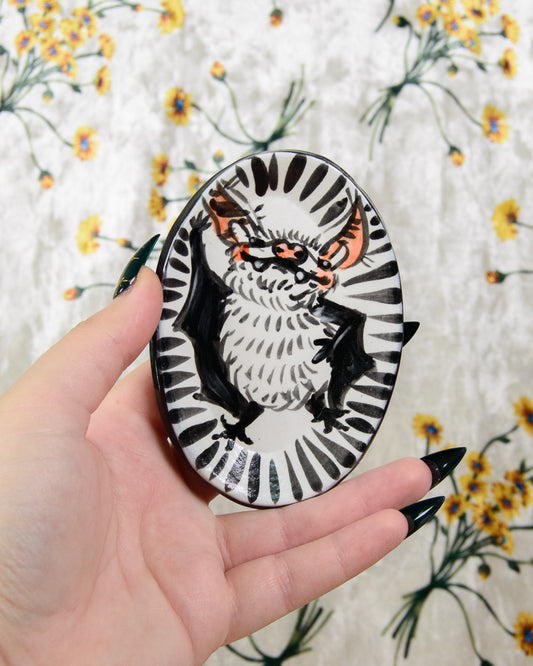 Hand holding a round ring dish with a bat design on a floral-patterned fabric background