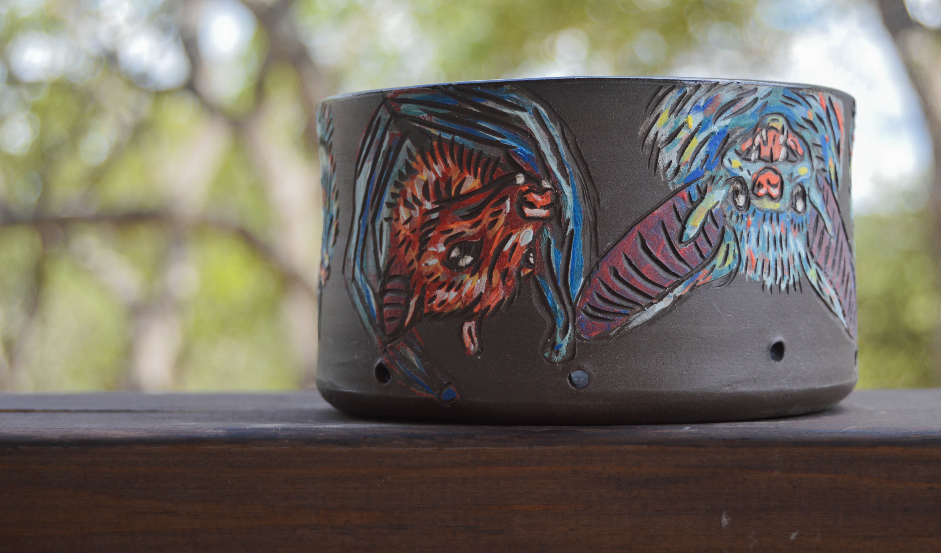 A handmade brown ceramic berry bowl with a pattern of bats around the edge, placed on a wooden surface with trees in the background.