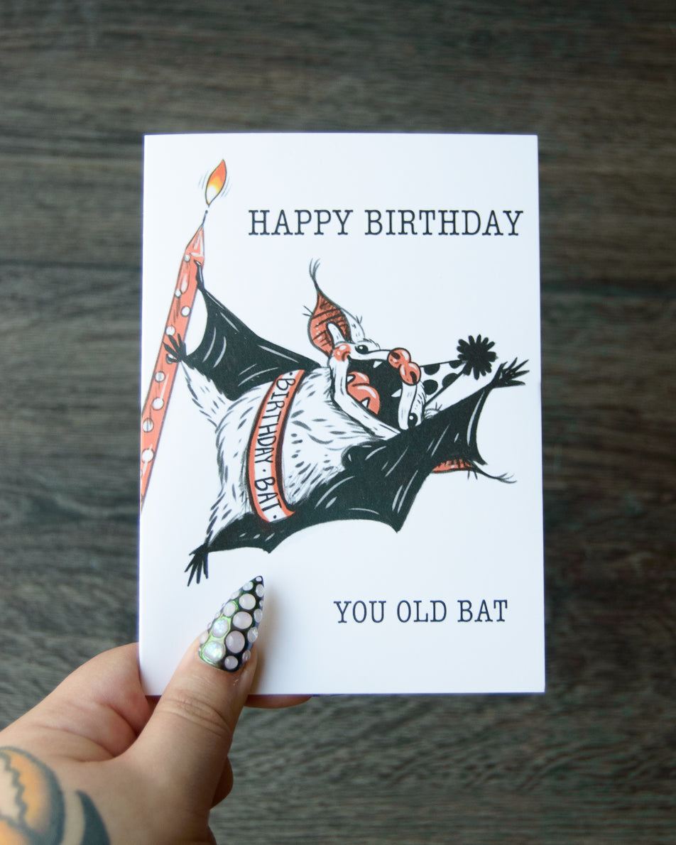 Birthday card with a bat illustration and text held by a hand on a wooden surface