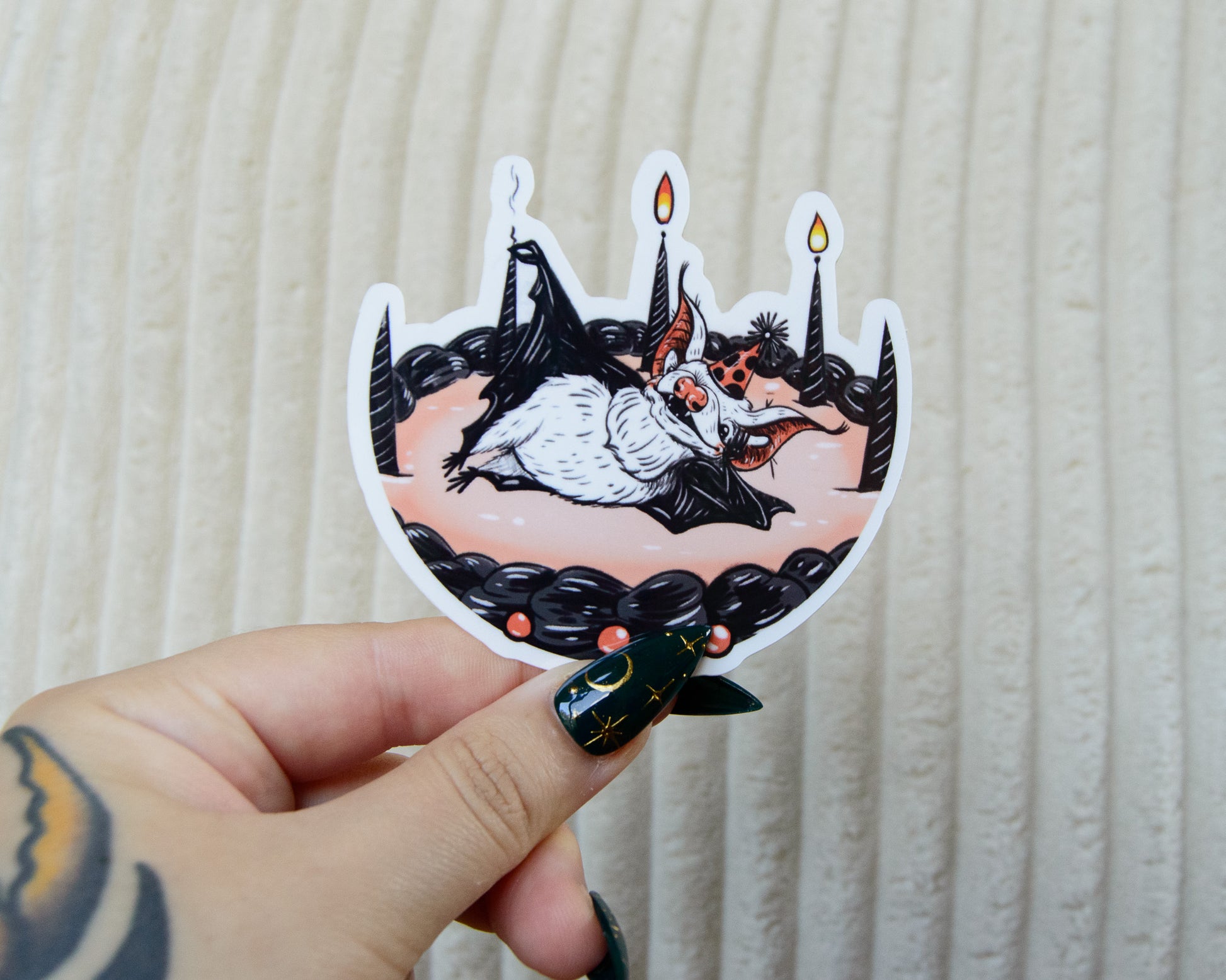 Hand holding a sticker with a design of a cute bat on a birthday cake with candles against a beige background
