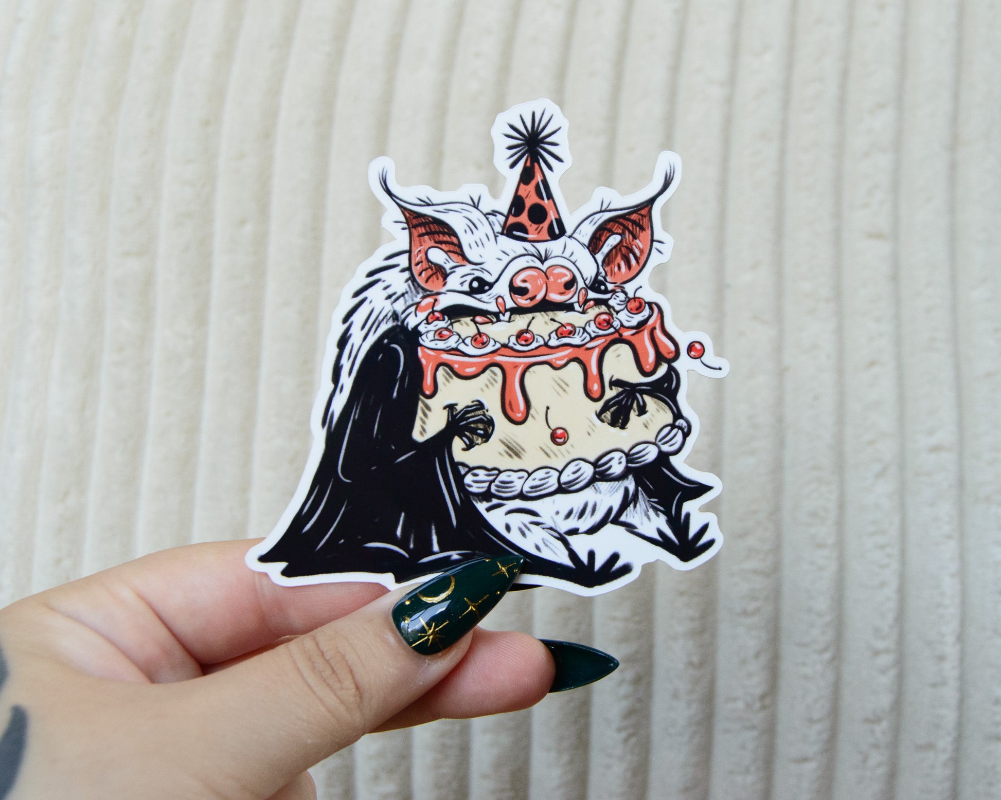 Hand holding a sticker with a design of a cute bat  in a party hat and eating a pink birthday cake against a textured beige background.