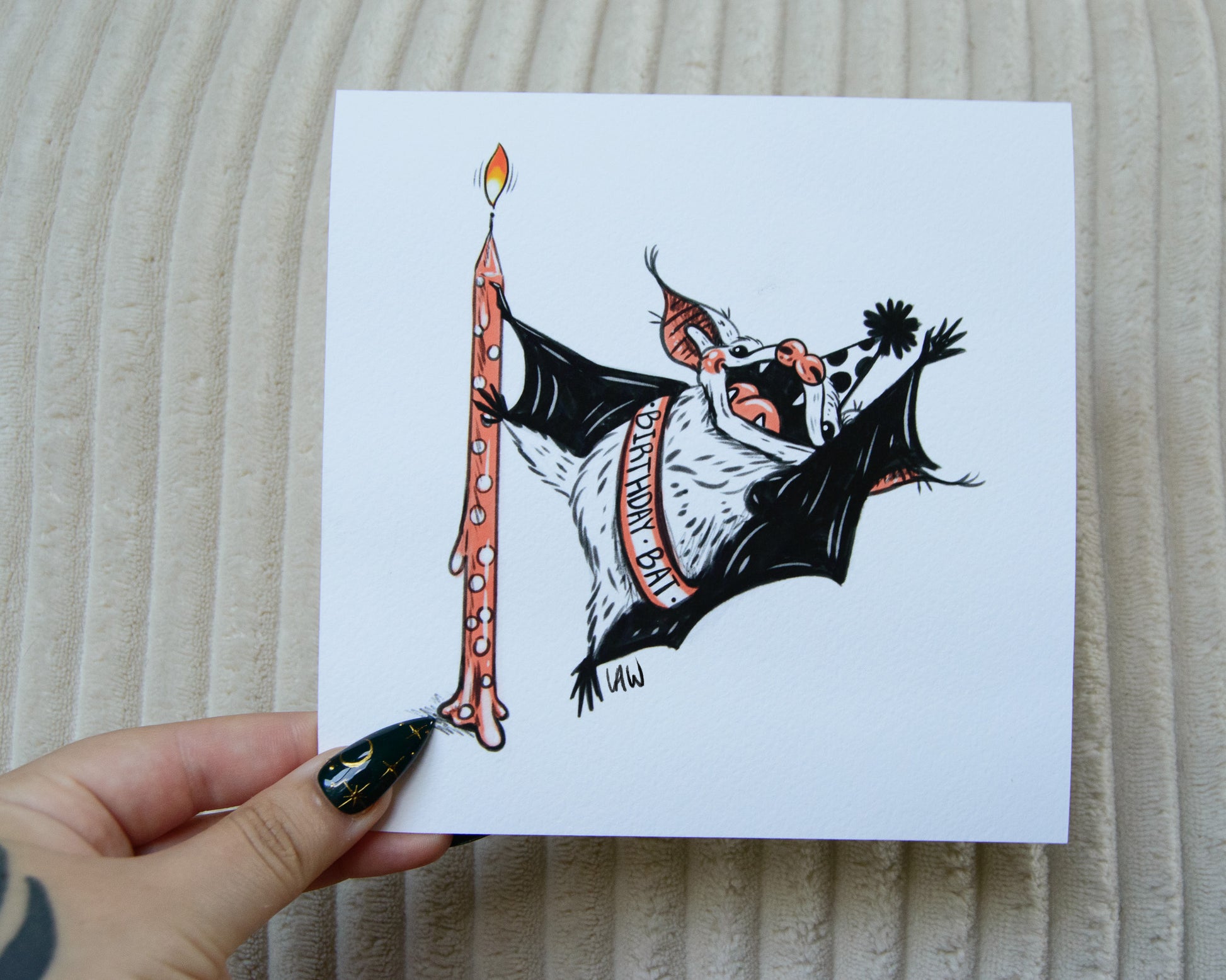 Illustration art print of a cute bat holding a birthday candle on a white background held by a hand against a textured beige background.