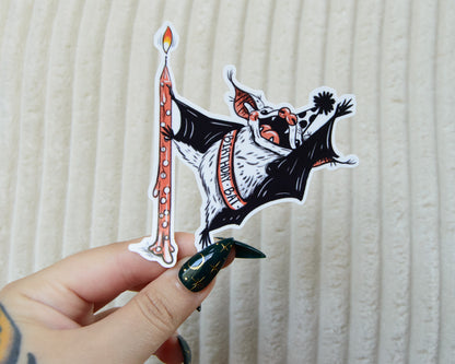 Sticker of a cute bat dancing around a birthday candle wearing a birthday sash and pointed party hat