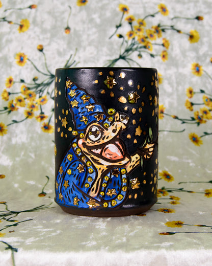 black ceramic cup with a cartoon frog wizard design and gold accents