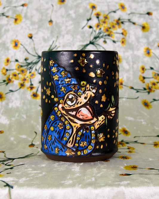 black ceramic cup with a cartoon frog wizard design and gold accents