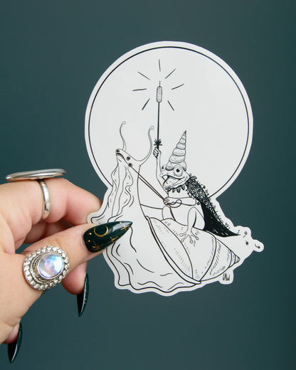 Hand holding a sticker with a black and white illustration of a frog riding a snail against a dark background.