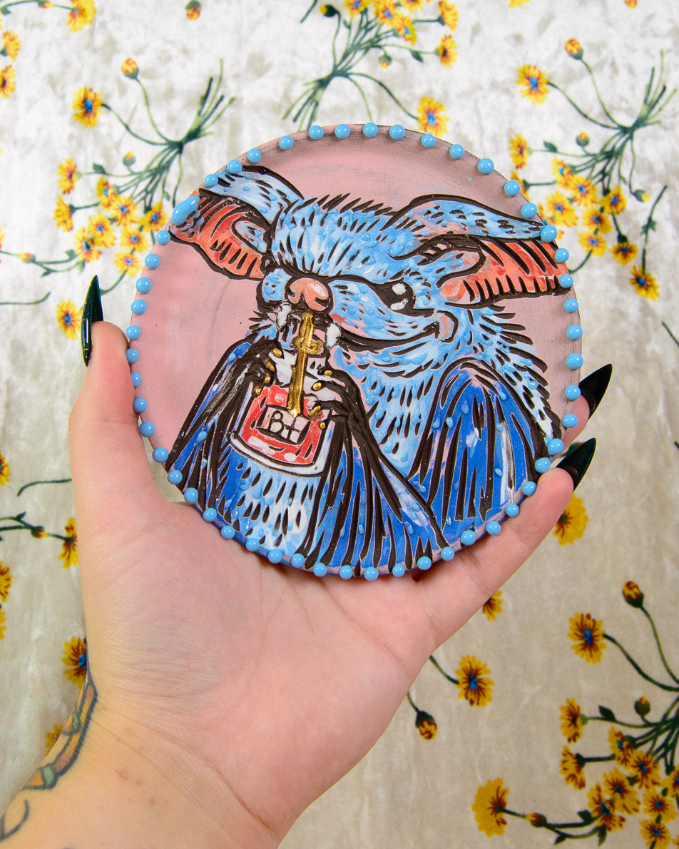 Coaster with a blue and black illustration of a bat holding a blood bag, held in a hand against a floral background.