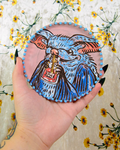 Coaster with a blue and black illustration of a bat holding a blood bag, held in a hand against a floral background.