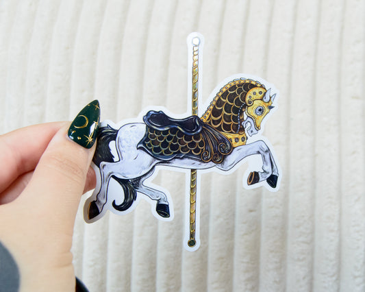 Hand holding a sticker of an intricate carousel horse illustration against a white background