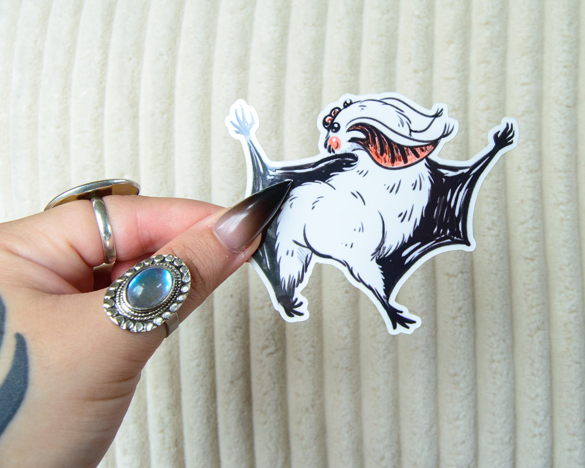 Sticker of a cartoon bat character with a white face and black wings against a beige background
