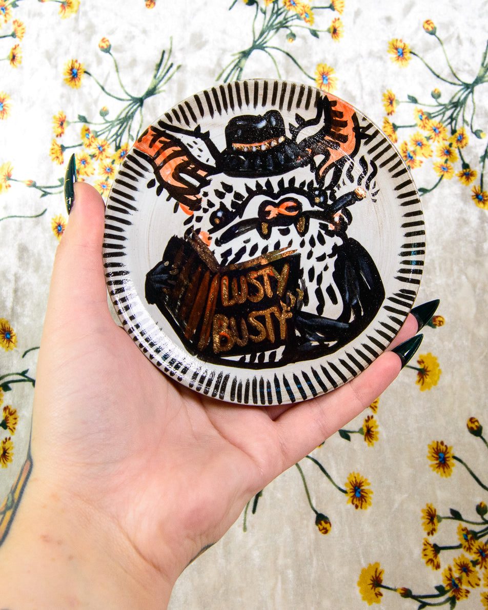 A black and white ceramic coaster with a cute cartoon design of a bat detective reading a book that says "lusty busty"