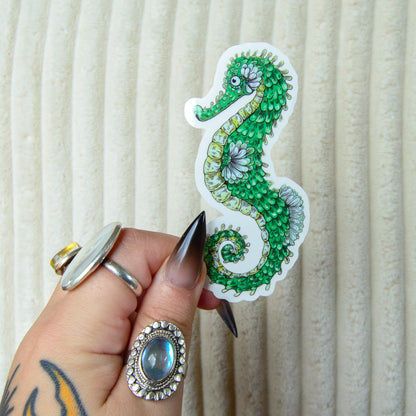 Hand holding a cute decorative green seahorse sticker against a beige background