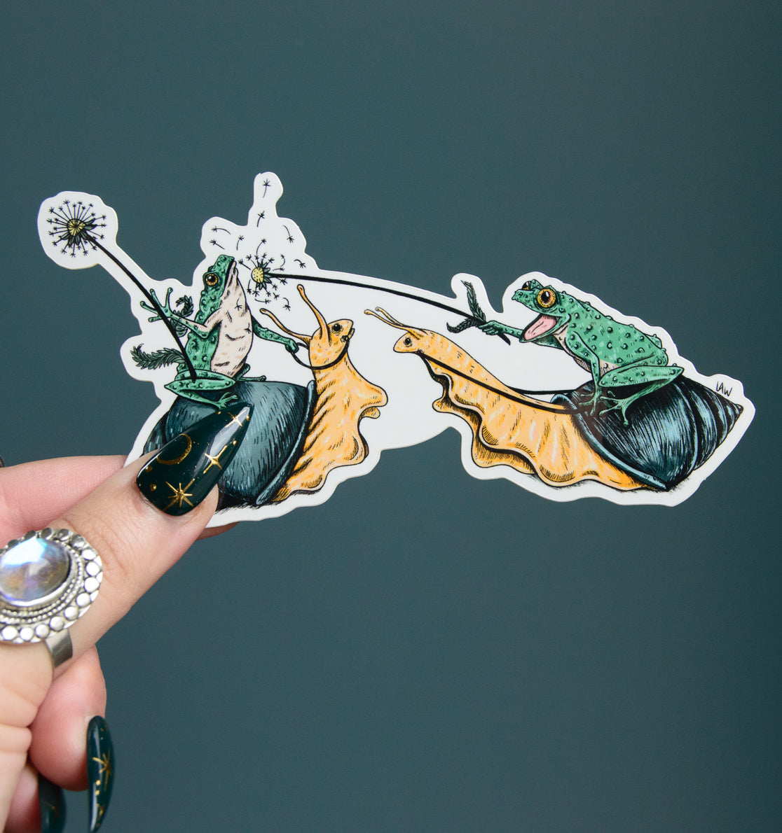 Hand holding a colorful sticker with frogs, jousting with dandelions and riding snails design against a dark background