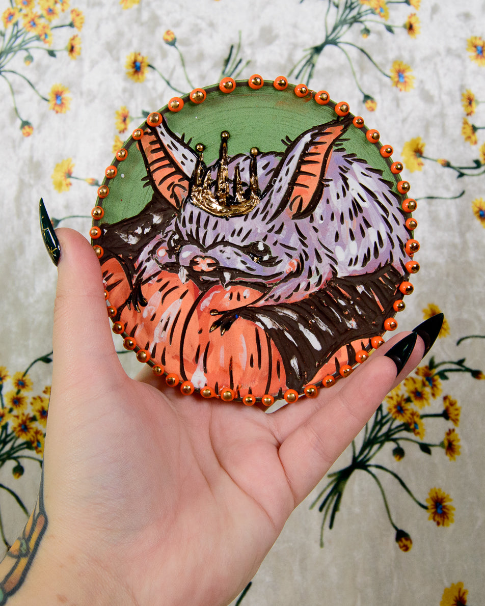 Decorative coaster with a colorful bat and pumpkin design held by a hand against a floral background