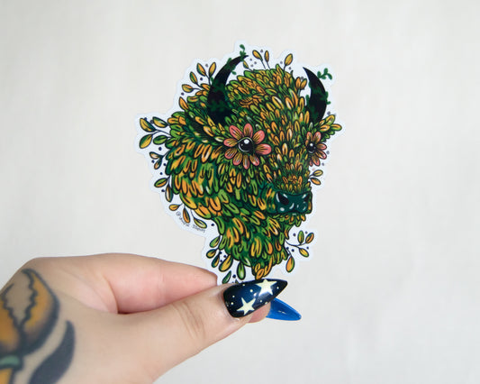 Hand holding a colorful illustrated sticker of a bison head made from leaves and flowers on a white background.