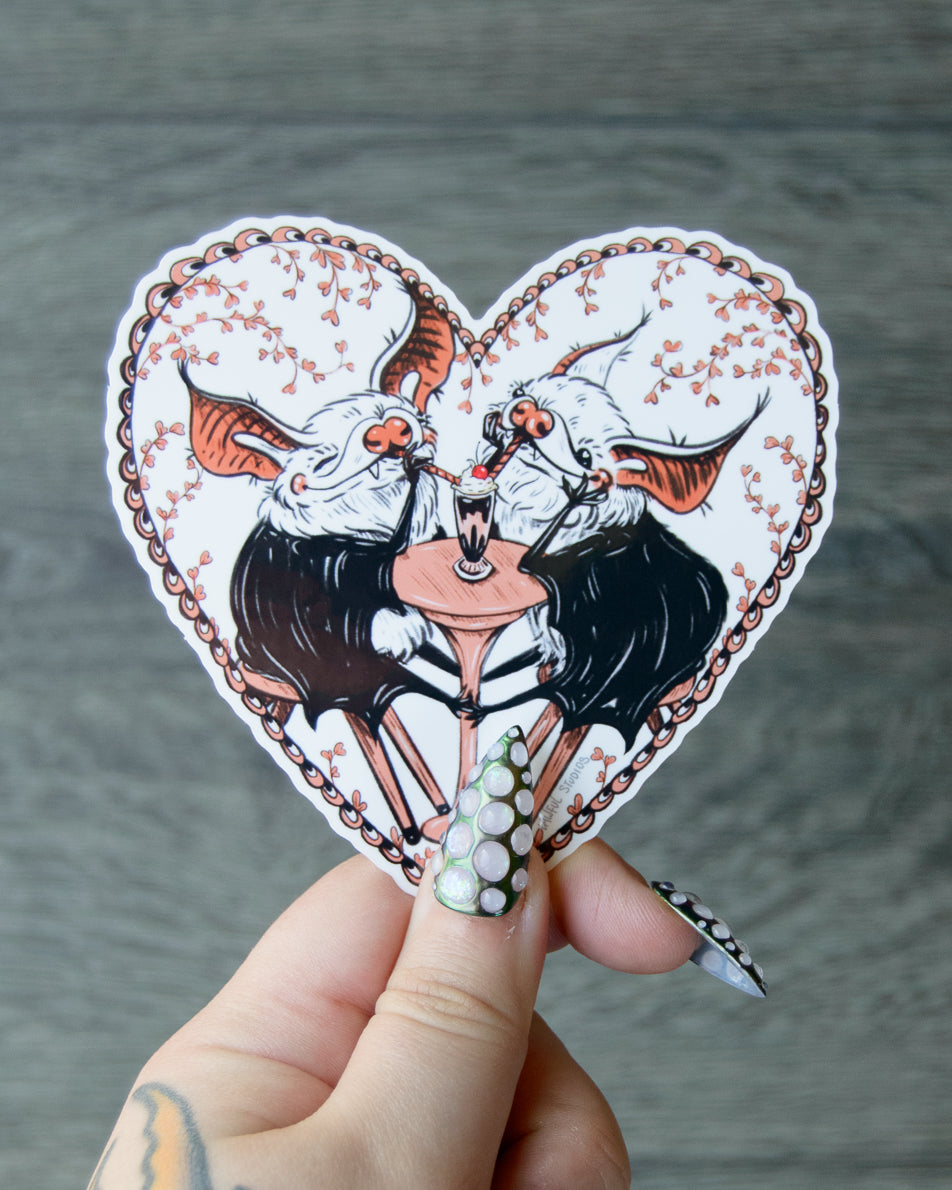 Heart-shaped sticker with two cartoon bats drinking from a milkshake held by a hand against a gray background