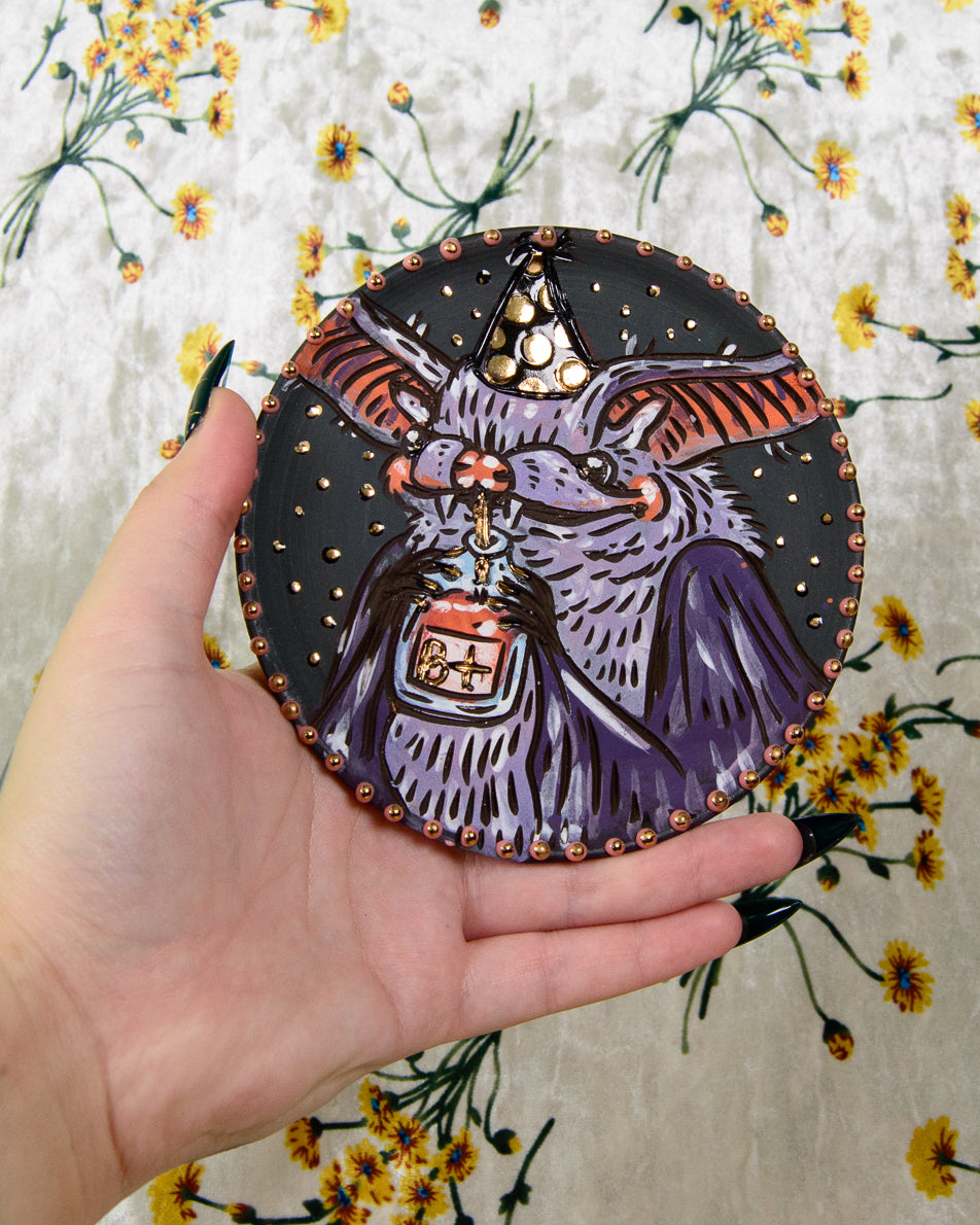 ceramic coaster with a cute purple bat wearing a party hat and drinking out of a B+ Blood bag
