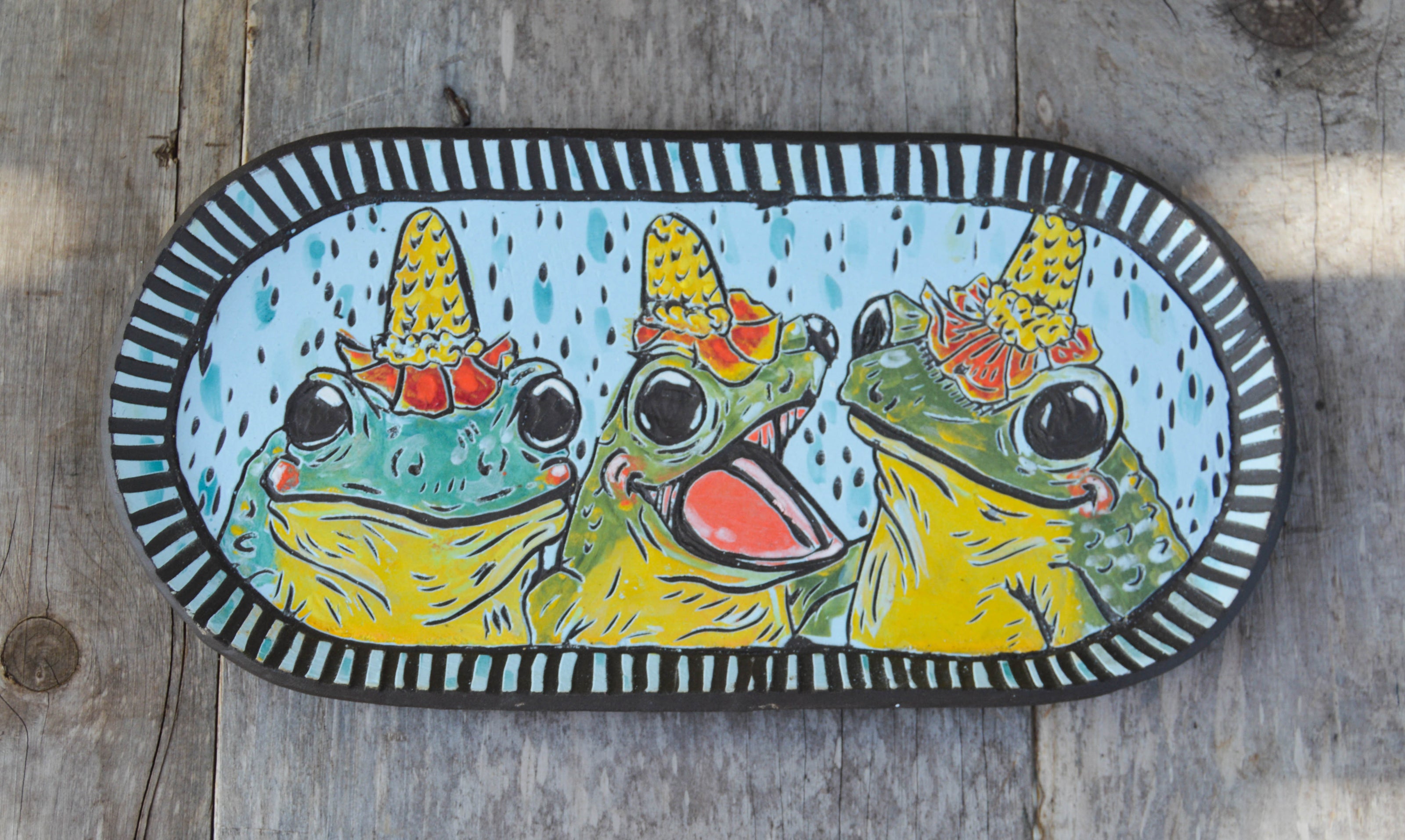 A decorative wall hanger featuring three illustrated frogs wearing flower hats on a wooden background.