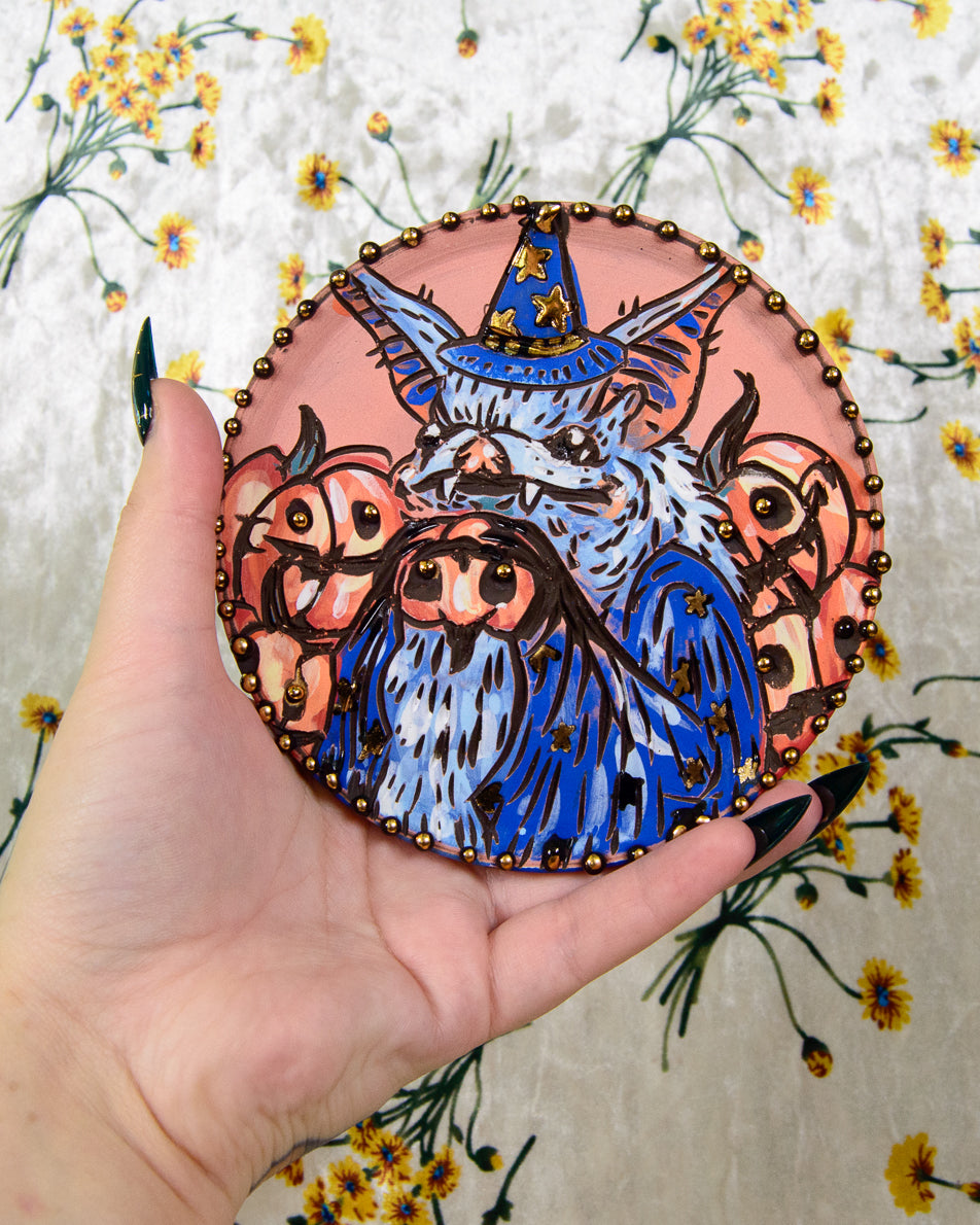 Hand holding a round, colorful ceramic coaster with a blue bat and pumpkin design against a floral background