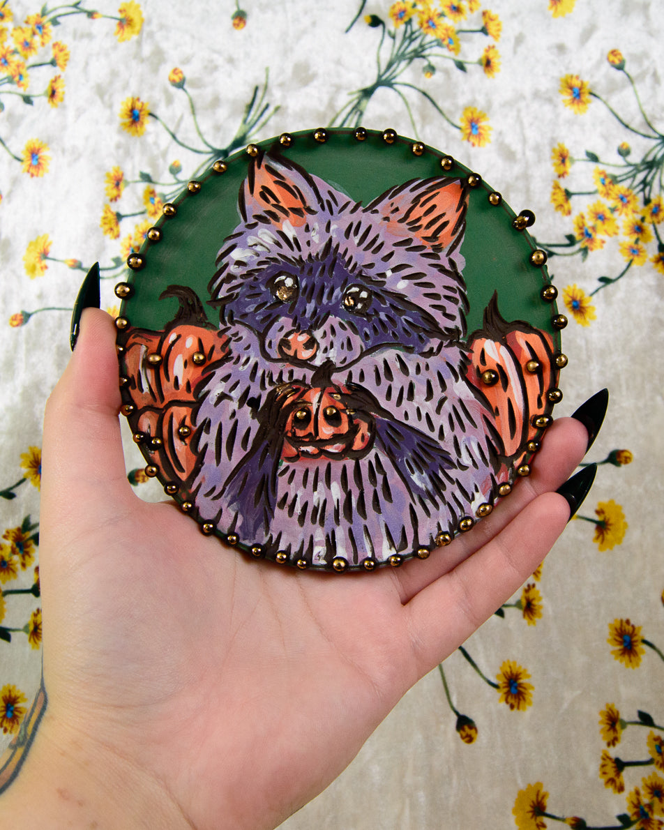 Hand holding a round, hand-painted ceramic coaster with a purple raccoon design against a halloween pumpkins