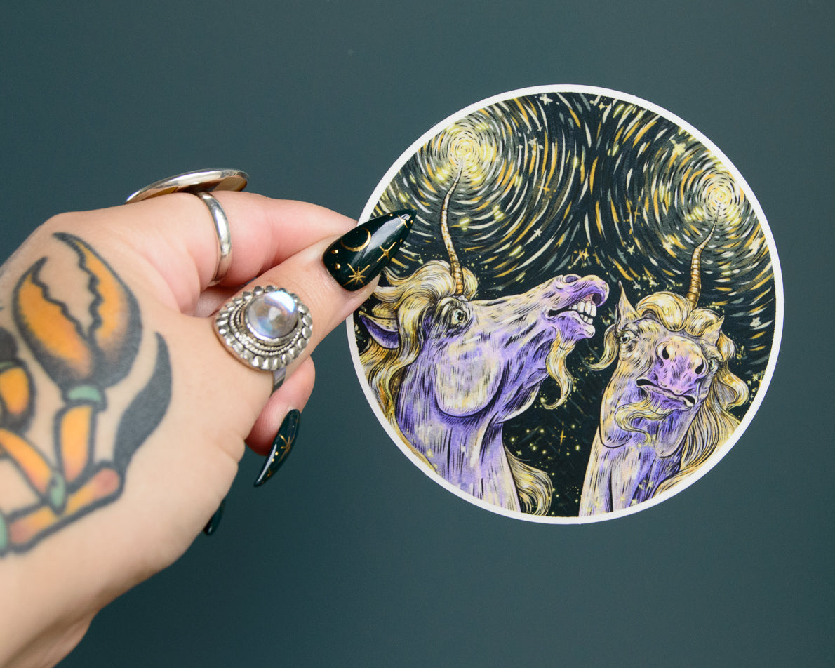 Hand holding a circular sticker with a stylized depiction of two unicorns against a starry night sky.