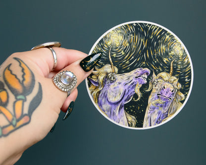 Hand holding a circular sticker with a stylized depiction of two unicorns against a starry night sky.
