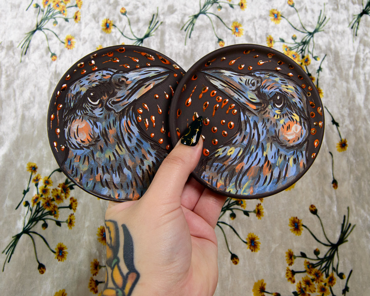 two round coasters with painted purple crow designs
