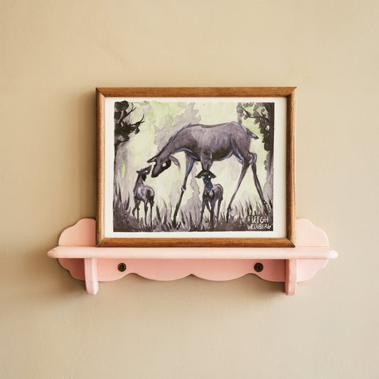 Framed artwork of deer, doe with baby fawns on a pink shelf against a beige wall