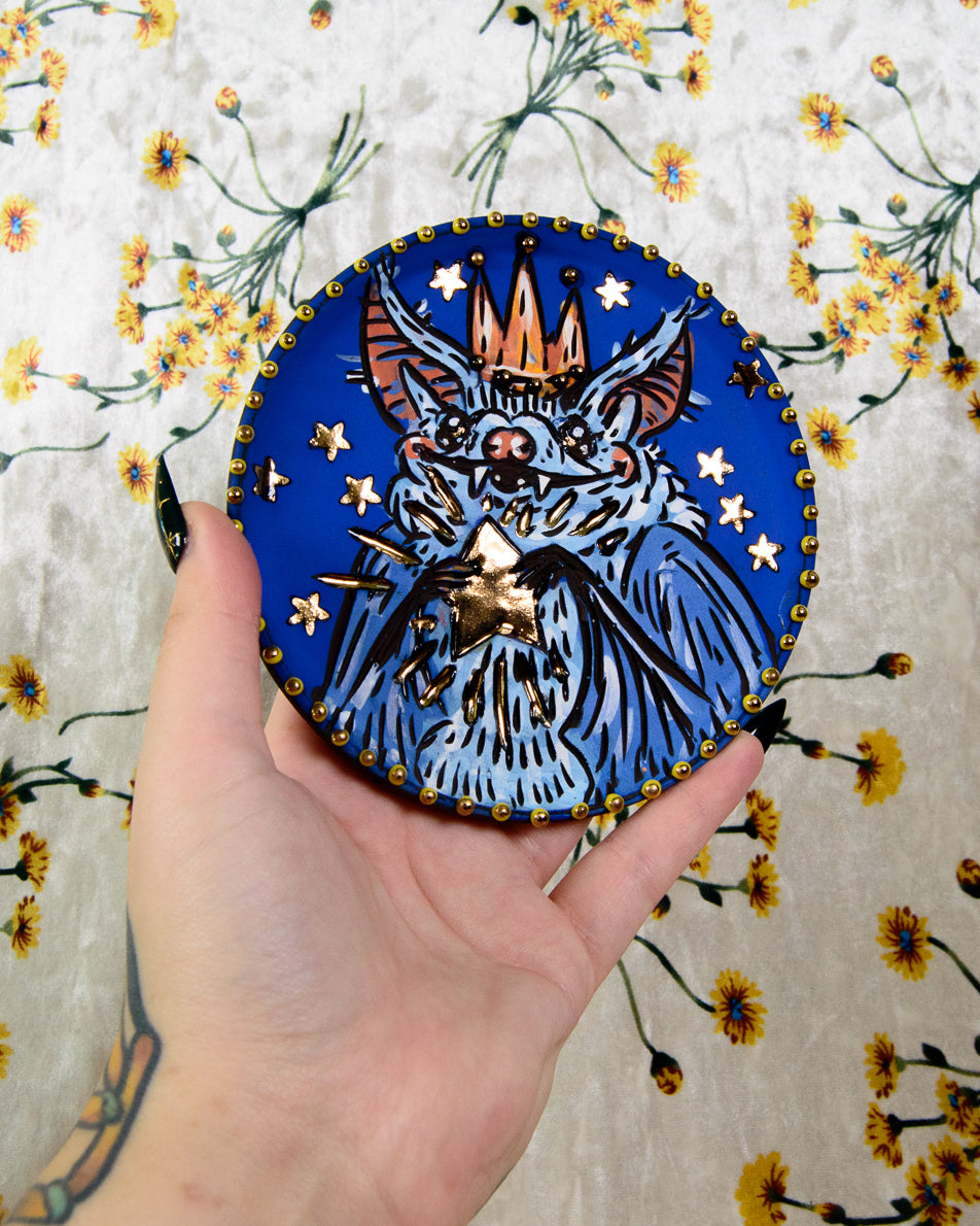 Handmade ceramic coaster with a colorful blue stylized bat holding golden stars