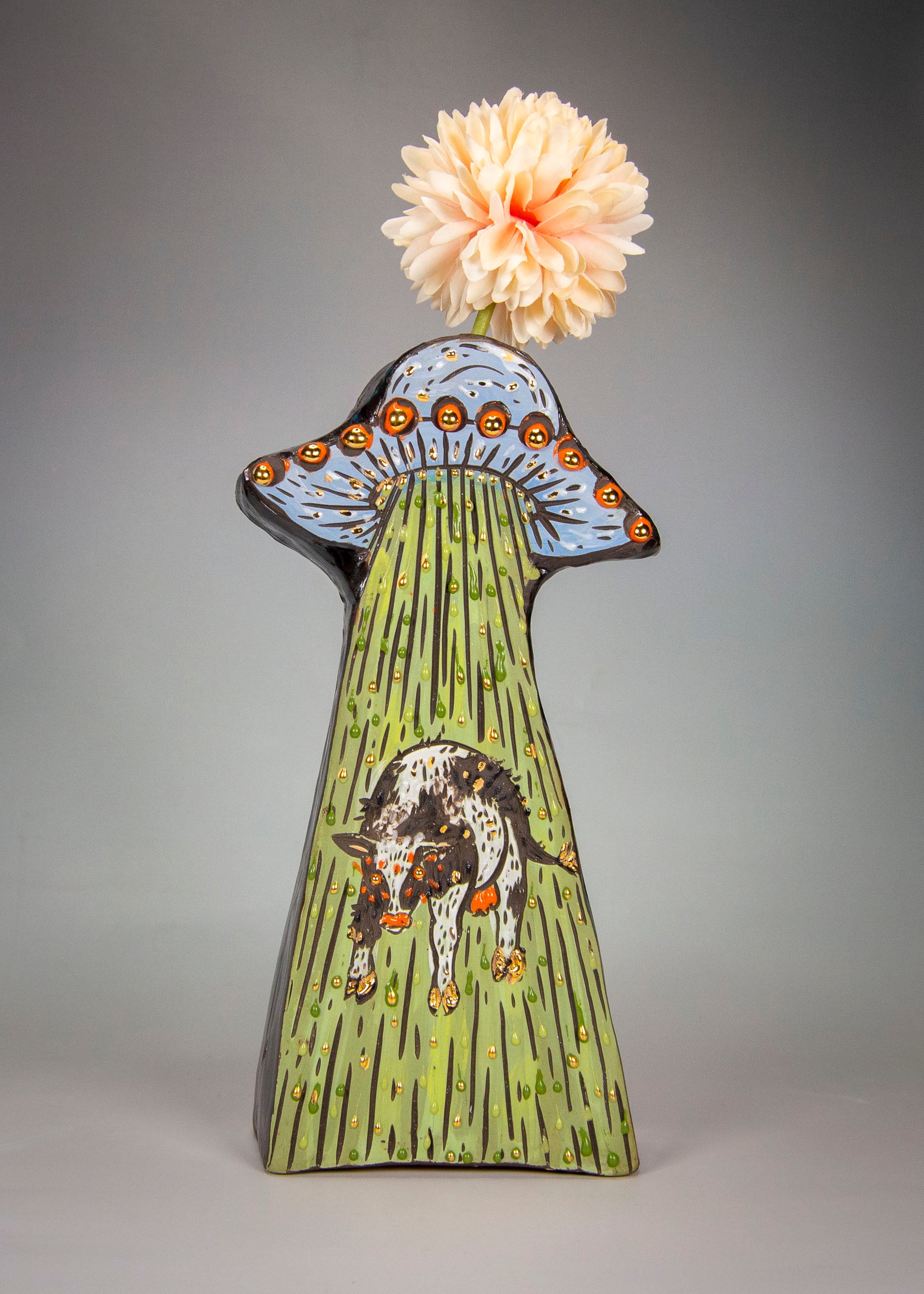 cartoonish stylized ceramic vase with a alien UFO and cow design with a flower on top against a gray background