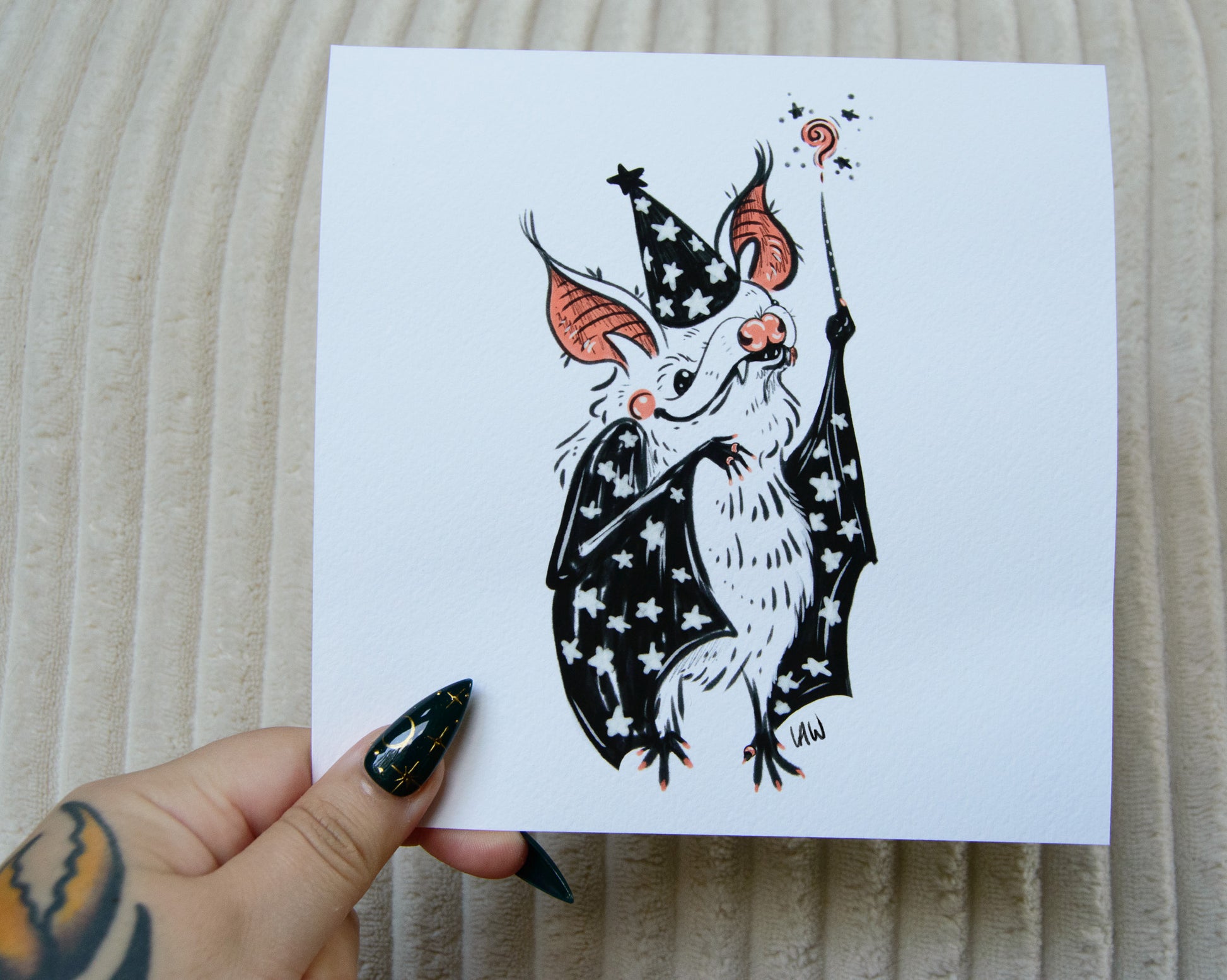 Hand holding a print of a black, pink, and white illustration of a bat wearing a witch or wizard hat and holding a magic wand