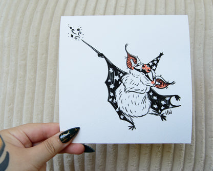 Hand holding a print with a black, pink, and white illustration of a bat wearing a witch / wizard hat on a beige textured background.